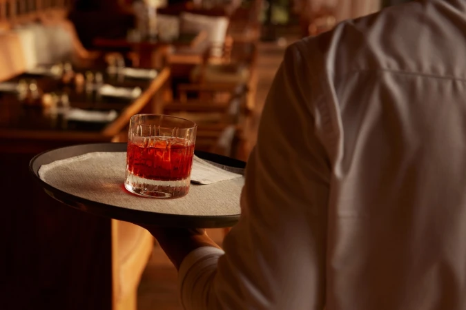 A waiter in a white shirt carries a cocktail glass filled with a red drink on a tray in a restaurant setting with warm lighting.