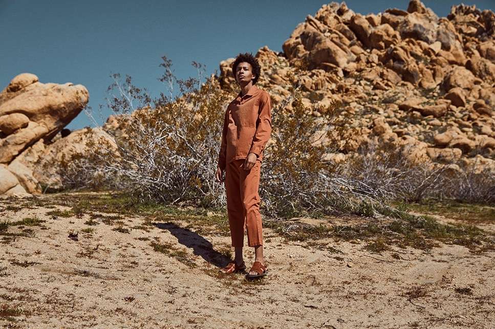 A person in matching shirt and pants stands in a desert landscape under a clear sky with sparse vegetation and rock formations.