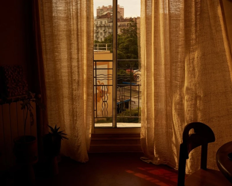 Indoor view through open balcony door with billowy burlap curtains, showing apartments, trees, and plants. Chair in foreground.