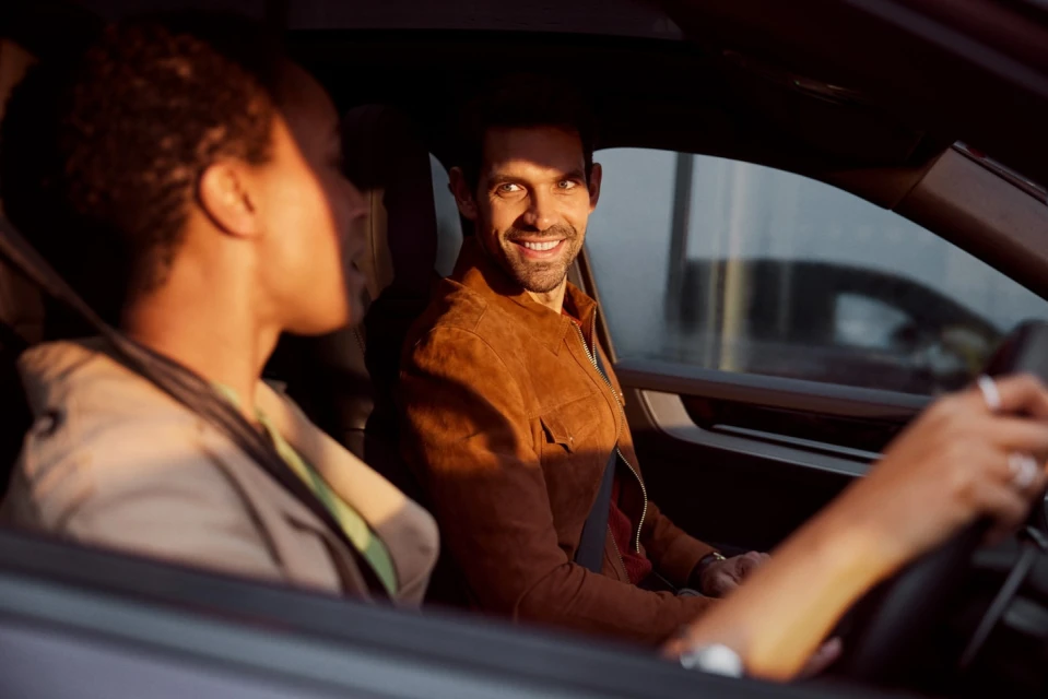 Couple in car interior, man in brown jacket smiling at driver, interior car shot, daytime lighting, modern vehicle.