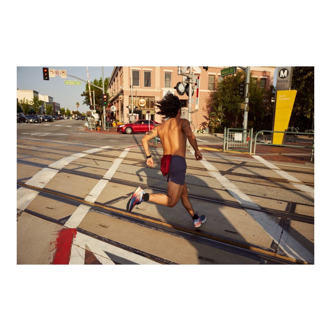 Shirtless man runs across train tracks in city street on sunny day wearing running shoes and fitness tracker with cars and buildings in the background.
