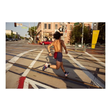 Shirtless man runs across train tracks in city street on sunny day wearing running shoes and fitness tracker with cars and buildings in the background.