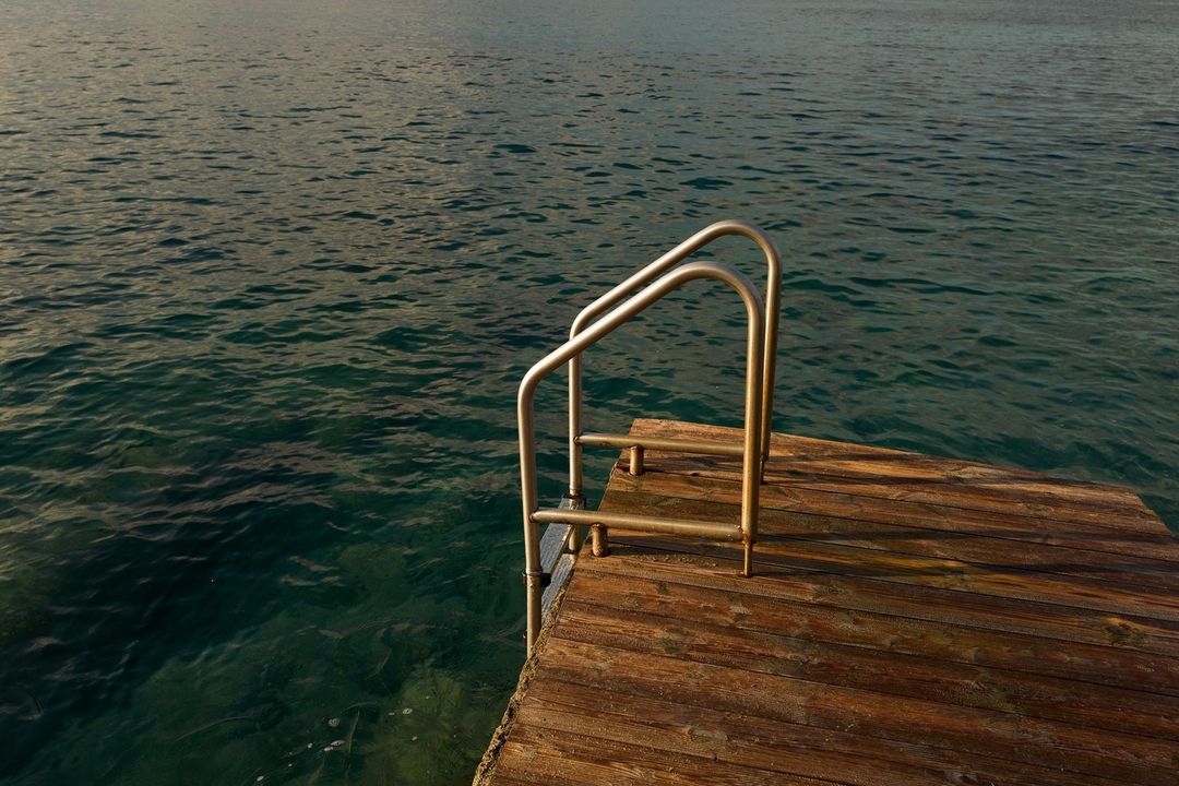 Wooden dock with a metal ladder leading into dark turquoise water, creating a serene scene of tranquility and a pathway to the sea.