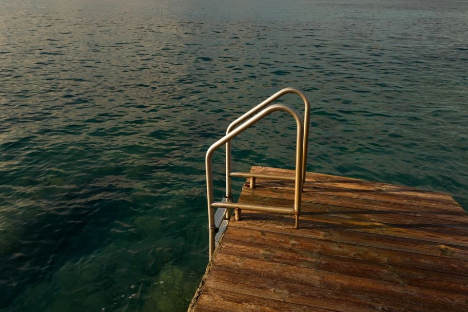 Wooden dock with a metal ladder leading into dark turquoise water, creating a serene scene of tranquility and a pathway to the sea.