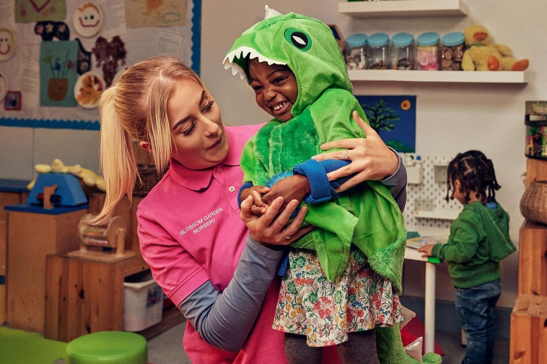 Teacher helping a child in a green dinosaur costume in a classroom with toys and learning materials in the background, creating a playful scene.