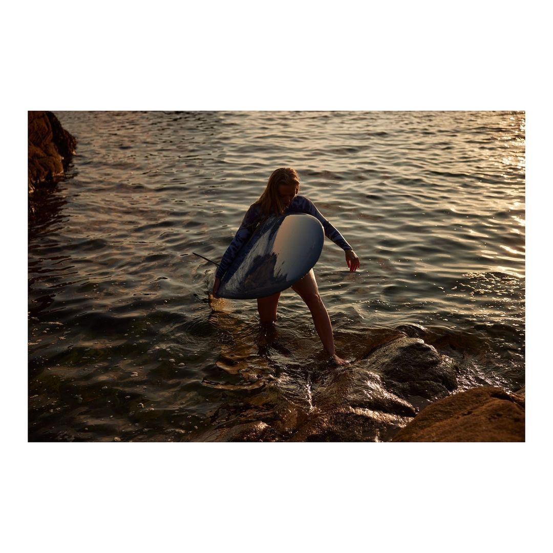 Woman in a wetsuit walks out of the ocean carrying a surfboard at sunset; rocks and golden light reflect in the water.