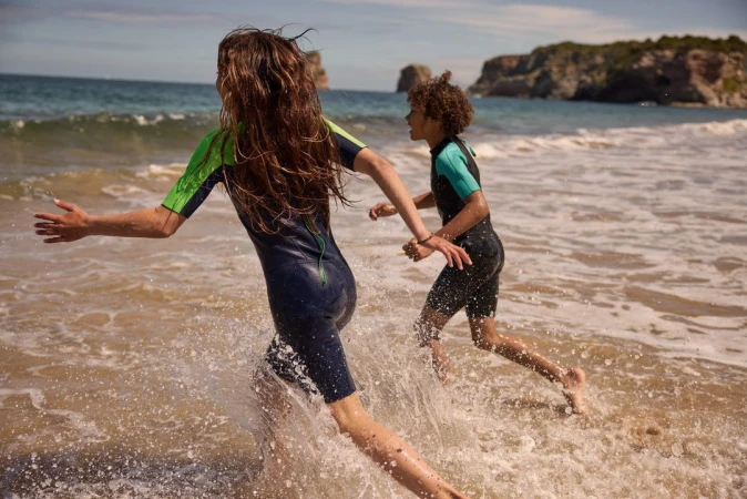 Two children in wetsuits run and splash in the surf at a beach with island formations and blue skies, enjoying a summer day.