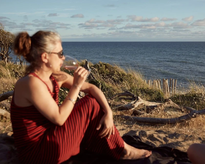 Woman in red striped dress drinks wine by the ocean at sunset. She sits on a blanket with beach vegetation in the background.