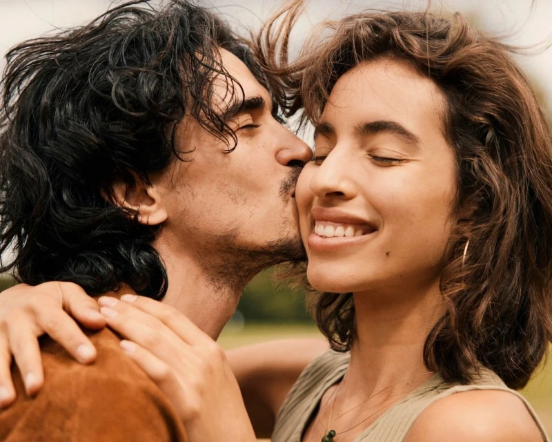 A smiling young woman with her eyes closed is being kissed on the cheek by a young man with long dark hair in an outdoor setting.