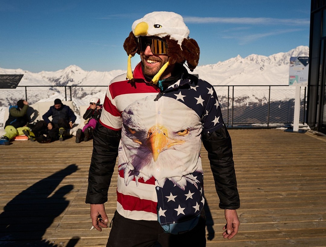 Man in eagle hat and American flag jacket smiles on ski resort deck with snow-covered mountains under a clear blue sky.