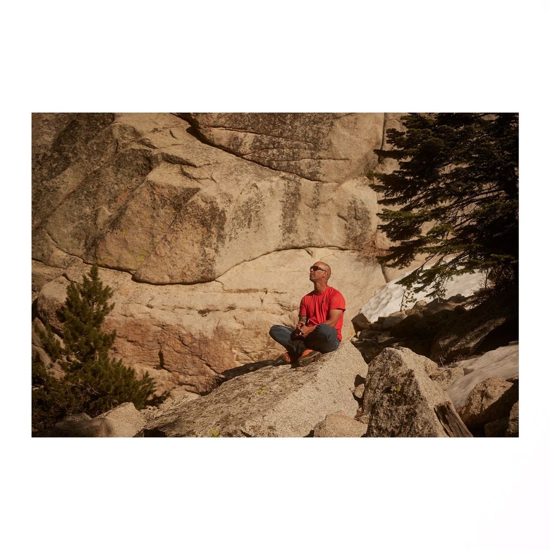 Adult male in red shirt and jeans sits cross-legged on a boulder outside, looking up at the stone cliffside with foliage.