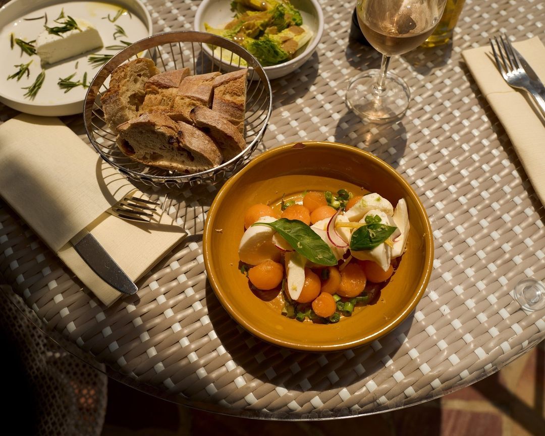 Overhead shot of a table with bread, salad, cheese, and wine, set on a woven surface in soft, natural light. 