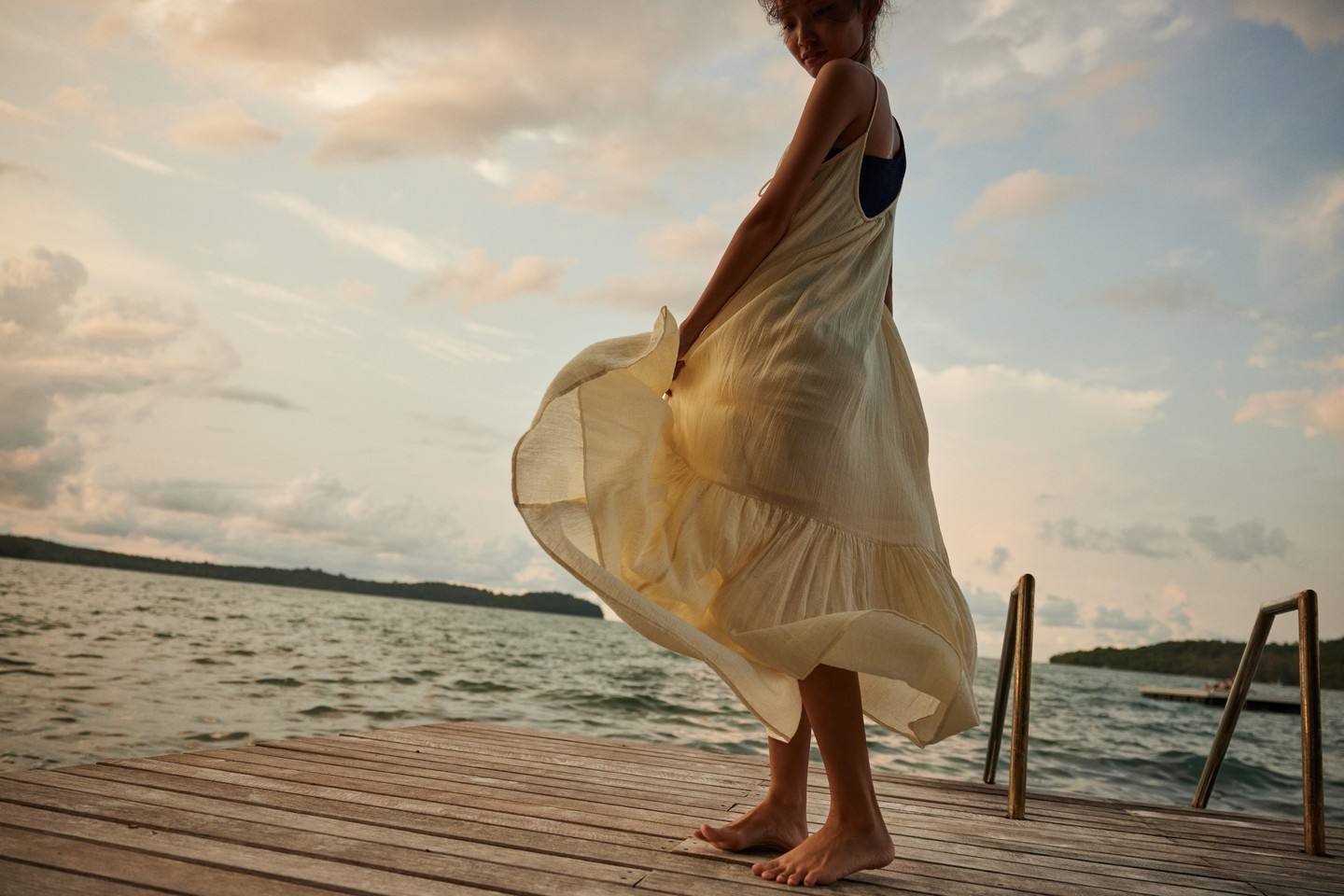 A woman in a flowing yellow dress stands on a wooden dock, enjoying the sunset by the water with a serene and carefree expression.