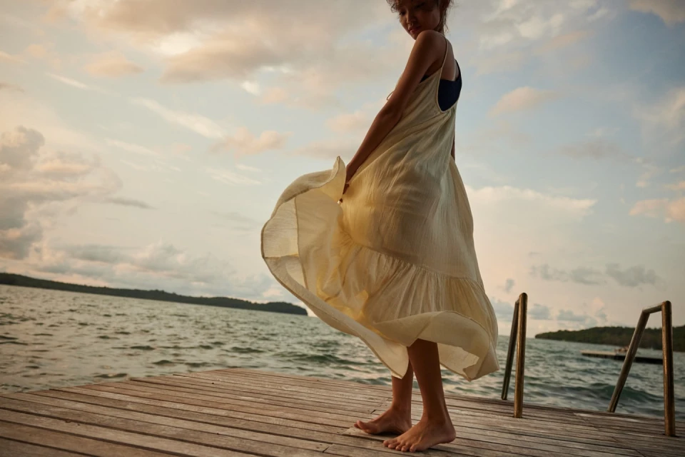 A woman in a flowing yellow dress stands on a wooden dock, enjoying the sunset by the water with a serene and carefree expression.