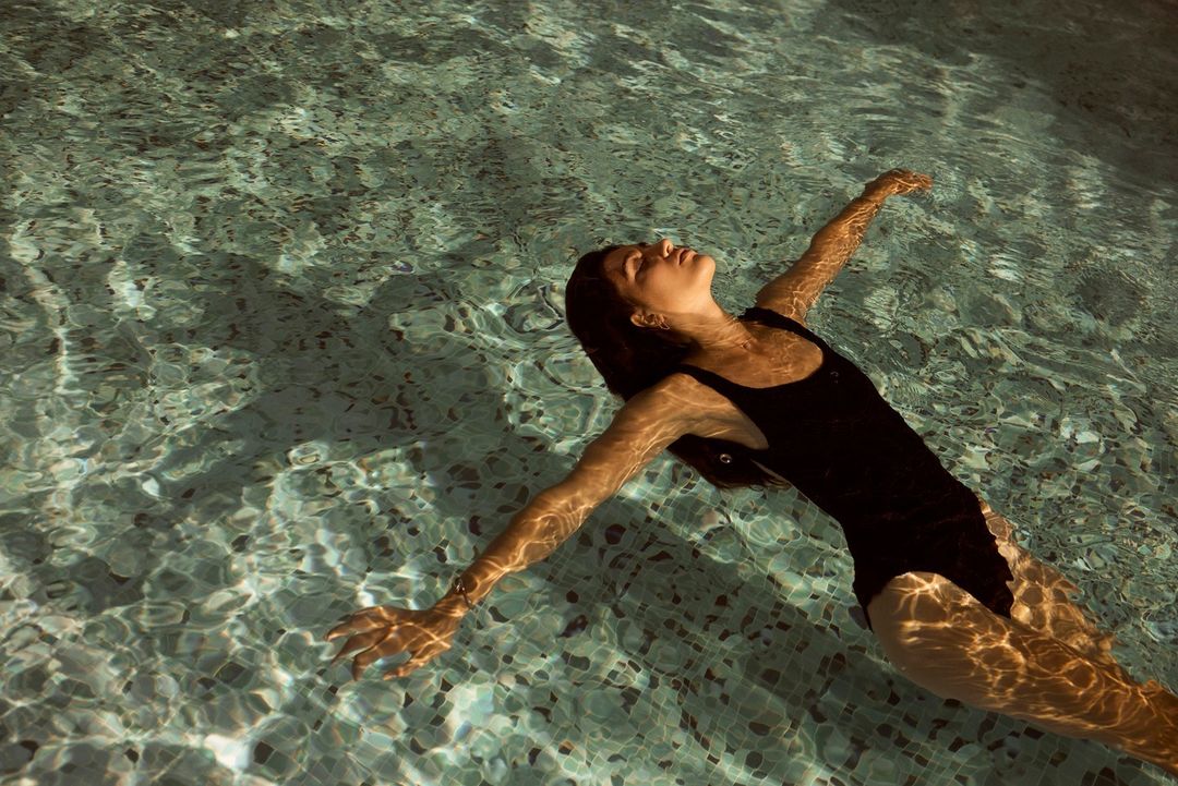 Woman in a black swimsuit floats in a pool with arms outstretched and head back, reflecting dappled sunlight and shadows on the water.