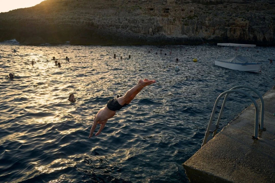 Man dives into ocean at sunset from concrete pier, with swimmers visible in the sea and a rocky cliff coastline in the background.