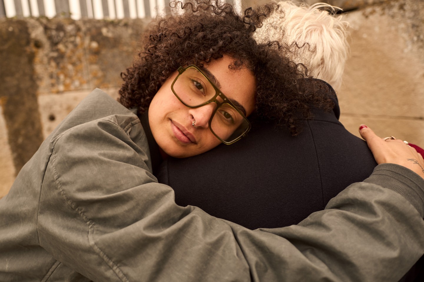 Two people embrace outdoors; one adult with curly hair and glasses smiles slightly; the other has light hair and dark coat.