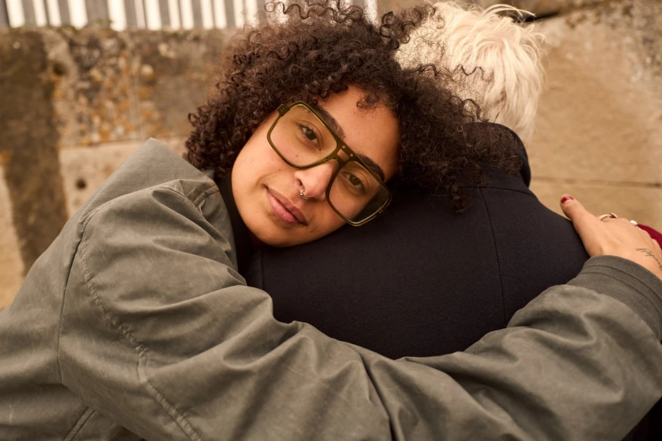 Two people embrace outdoors; one adult with curly hair and glasses smiles slightly; the other has light hair and dark coat.
