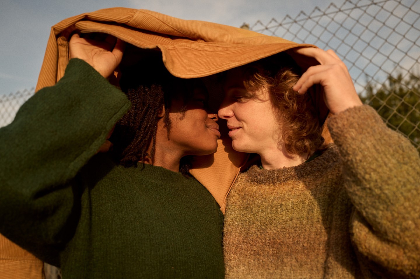 Two adults under a tan jacket near a chain-link fence. One adult with locs is on the left, while the other has curly blonde hair. They appear to be a couple.