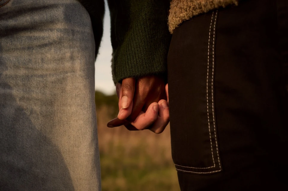 Close-up of two individuals of different skin tones holding hands outdoors, showing details of their hands and lower torsos against a blurred grassy background.