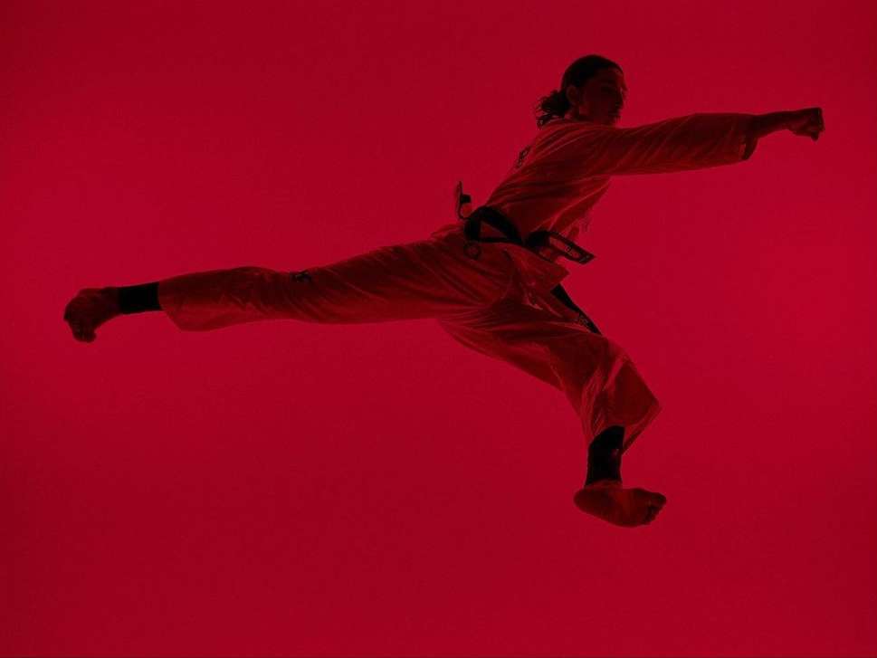 A martial artist in mid-air executing a punch, wearing a white gi with a black belt, set against a saturated red backdrop.