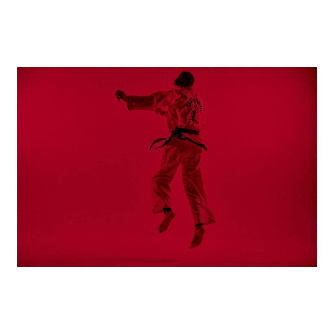 A person in a martial arts uniform is jumping, in a studio setting with red lighting. They are performing a kick in mid-air.