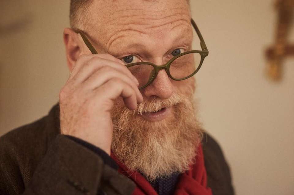 Close-up portrait of a red-bearded man with blue eyes wearing glasses, a dark jacket and a red scarf in an indoor setting.