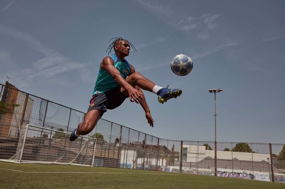 Athlete in mid-air kicking a soccer ball on a green field under a blue sky, buildings and fencing in background. Sports photography.