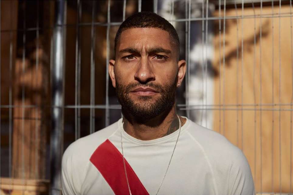 Portrait of a handsome adult male with a beard, wearing a white shirt with a red stripe, with a metal fence in the background.