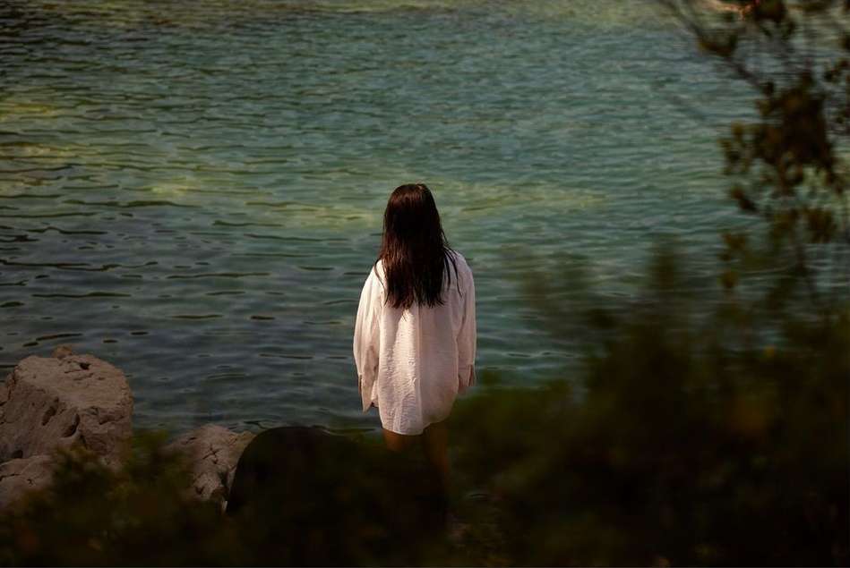 A woman stands on a rocky shore looking out to a calm body of water surrounded by vegetation in the background. 