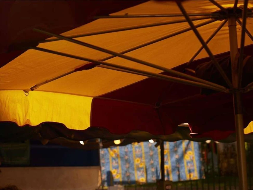 Patio umbrella with yellow and red fabric and metal frame, providing shade over an outdoor setting, with a glimpse of patterned fabric.
