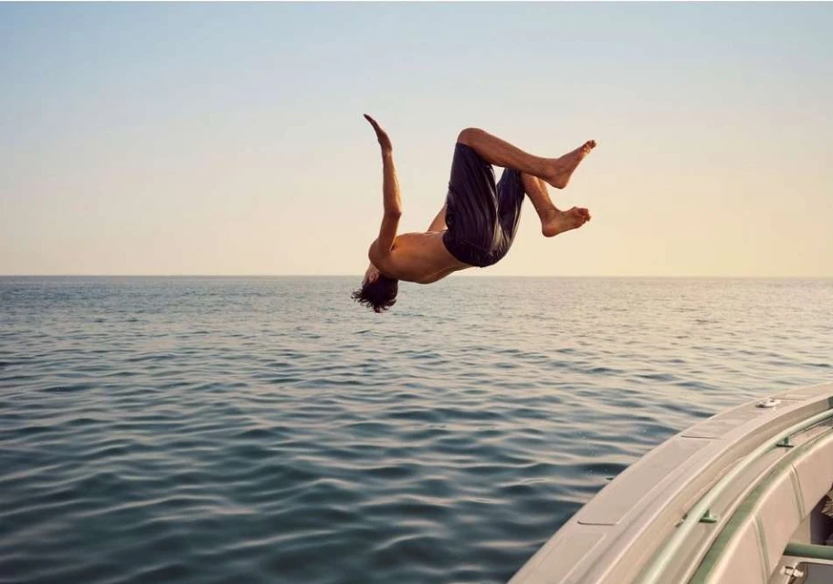 Athletic adult male does a mid-air backflip over the ocean, wearing swim trunks, boat visible in foreground, horizon clear.