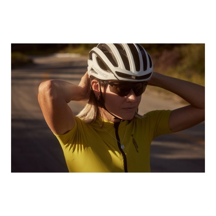 Woman cyclist adjusts helmet outdoors on a sunny day. She wears sunglasses and a yellow jersey. The road is visible in the background.