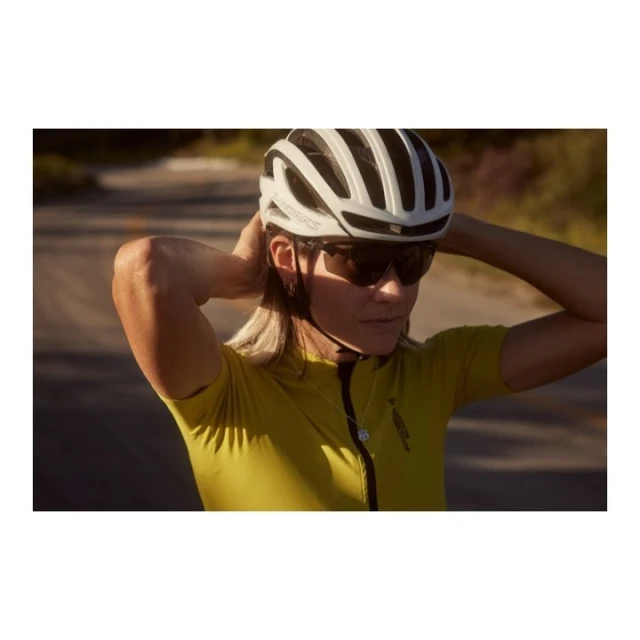 Woman cyclist adjusts helmet outdoors on a sunny day. She wears sunglasses and a yellow jersey. The road is visible in the background.