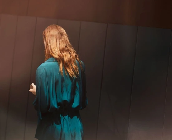 Rear view of a woman with long red hair wearing a teal silk shirt standing against a dark panelled wall