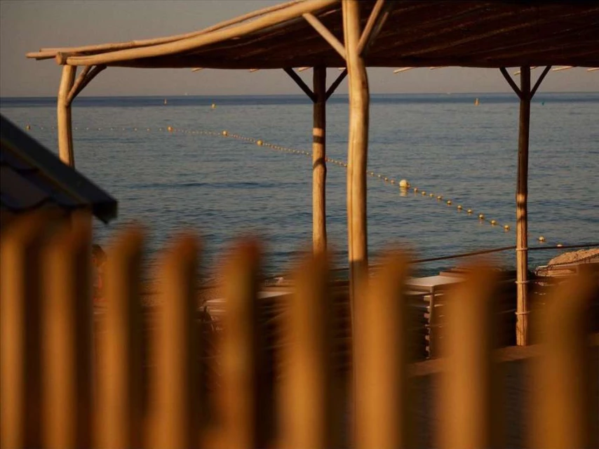 Seaside view through a blurred wooden fence of a beach shelter and calm ocean with a string of buoys under a light blue sky.