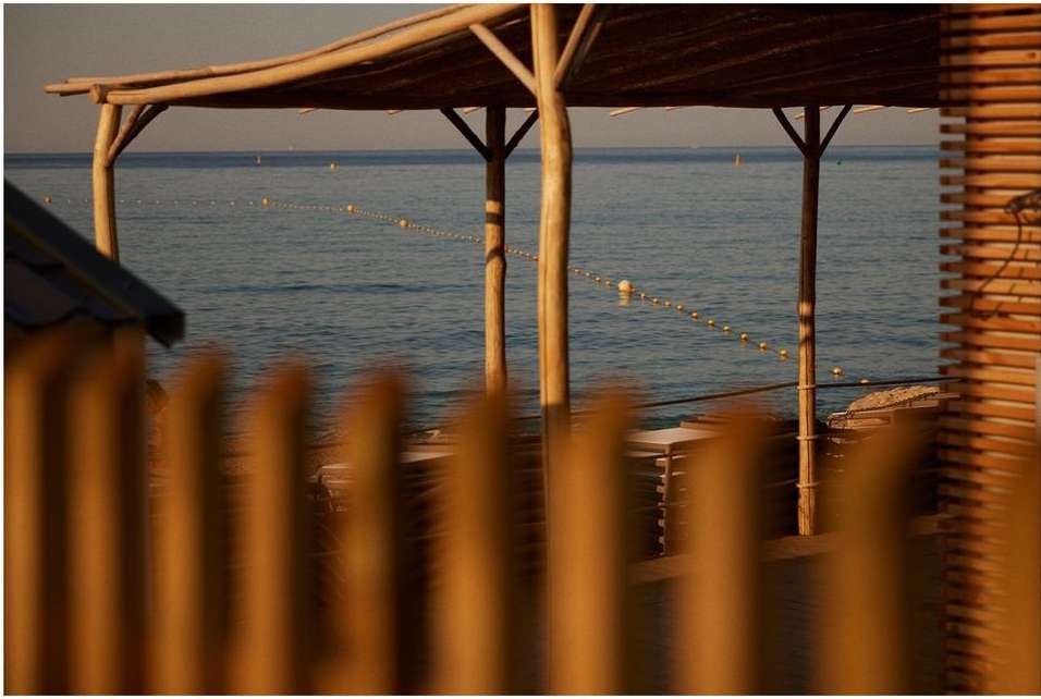 Seascape view through wood structure featuring beach umbrellas, calm ocean, and horizon on a sunny day at a coastal resort.