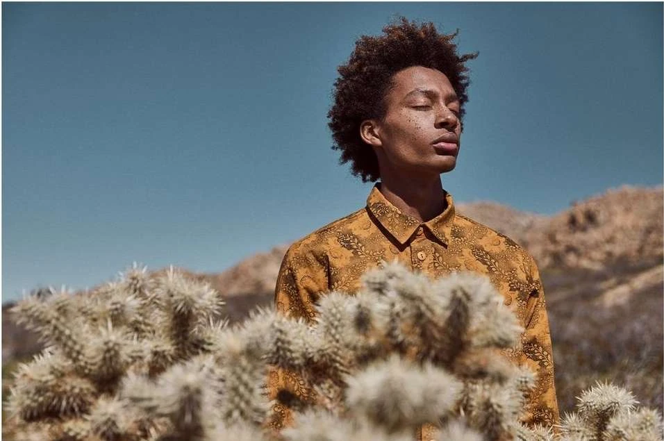 Portrait of a young adult male with curly hair and a patterned shirt, standing in front of desert vegetation under a clear blue sky.