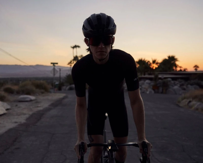 Cyclist in black riding a bicycle on an asphalt road at sunset with a mountain and palm trees in the background, wearing a helmet and sunglasses.