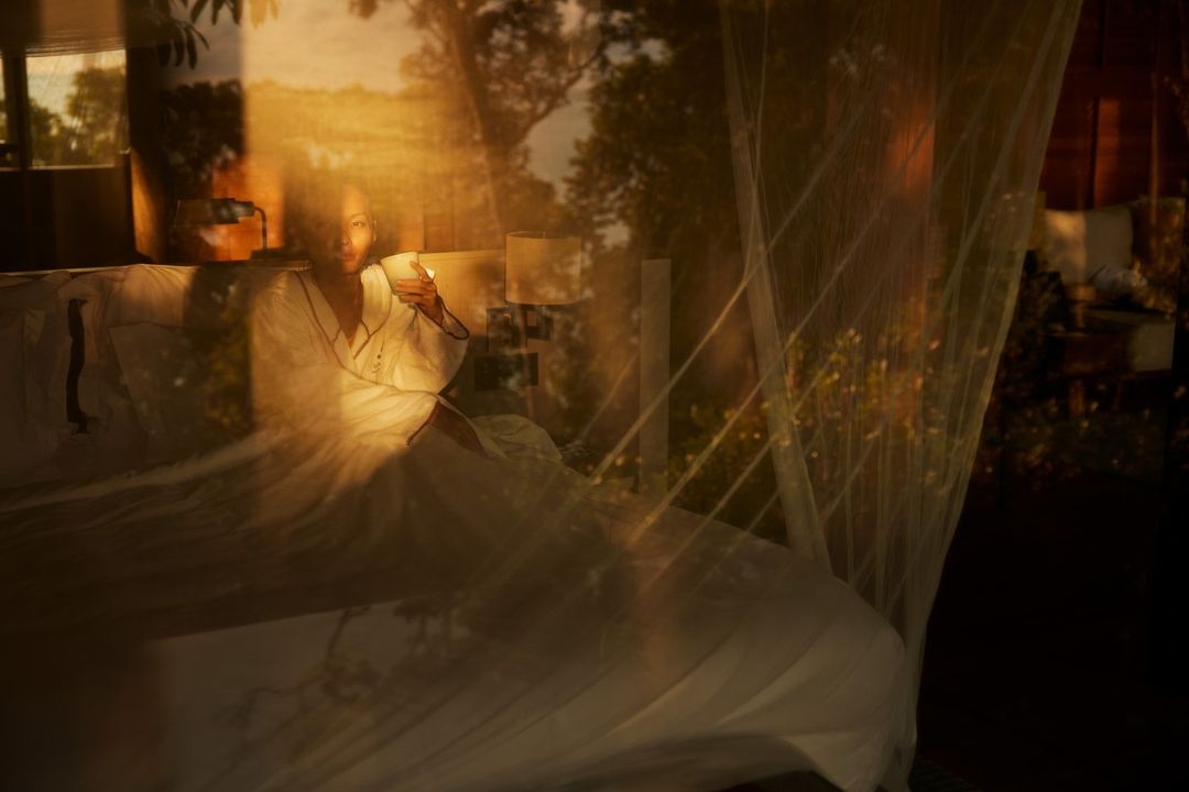 Woman in white robe sits in bed behind mosquito net, holding a cup, bathed in golden light, trees visible through a window, serene setting.