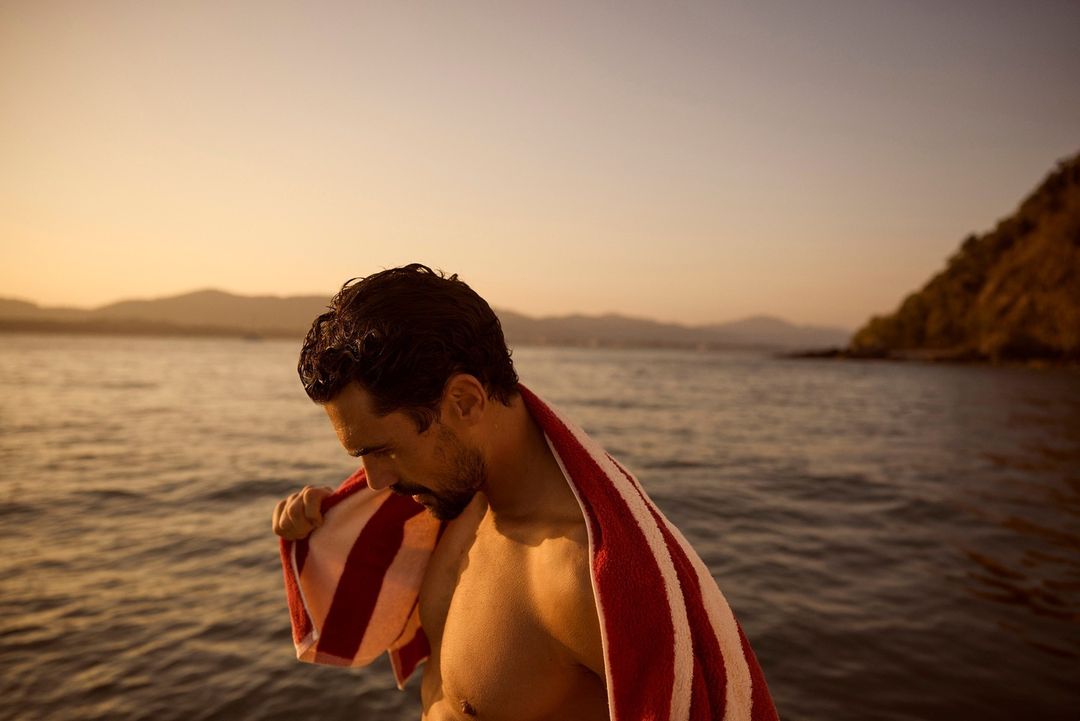 Man with dark hair and a towel stands on a beach near the ocean at sunset, with mountains visible in the background under a golden sky.