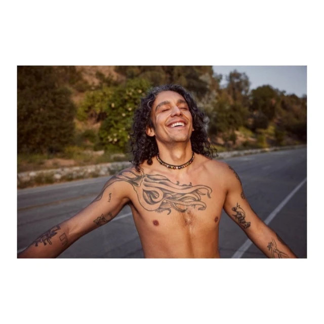 Smiling man with tattoos and long curly hair stands shirtless on a road in a rural setting on a bright day.