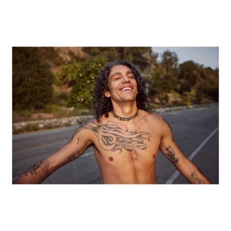 Smiling man with tattoos and long curly hair stands shirtless on a road in a rural setting on a bright day.