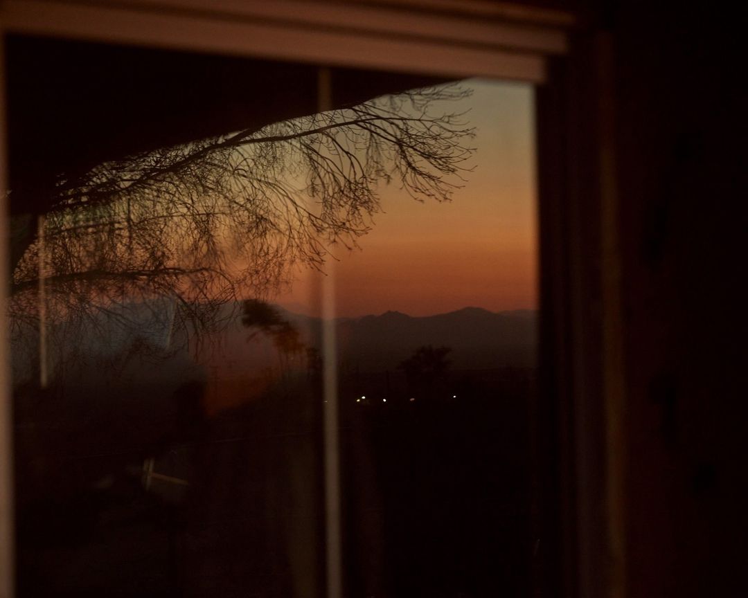 Window view of bare tree branches silhouetted against orange sunset sky, with distant mountain range in background.