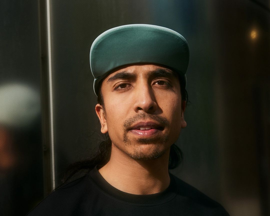 Portrait of a man with dark hair wearing a green cap and black shirt, looking at the camera with a neutral expression.
