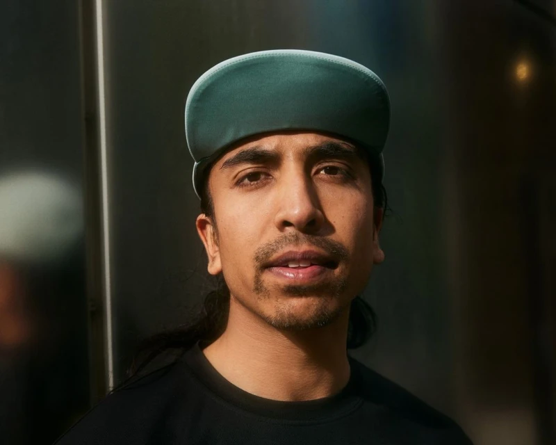 Portrait of a man with dark hair wearing a green cap and black shirt, looking at the camera with a neutral expression.