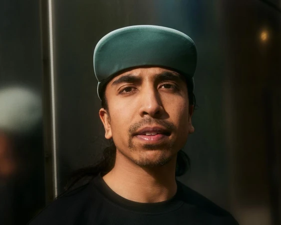 Portrait of a man with dark hair wearing a green cap and black shirt, looking at the camera with a neutral expression.