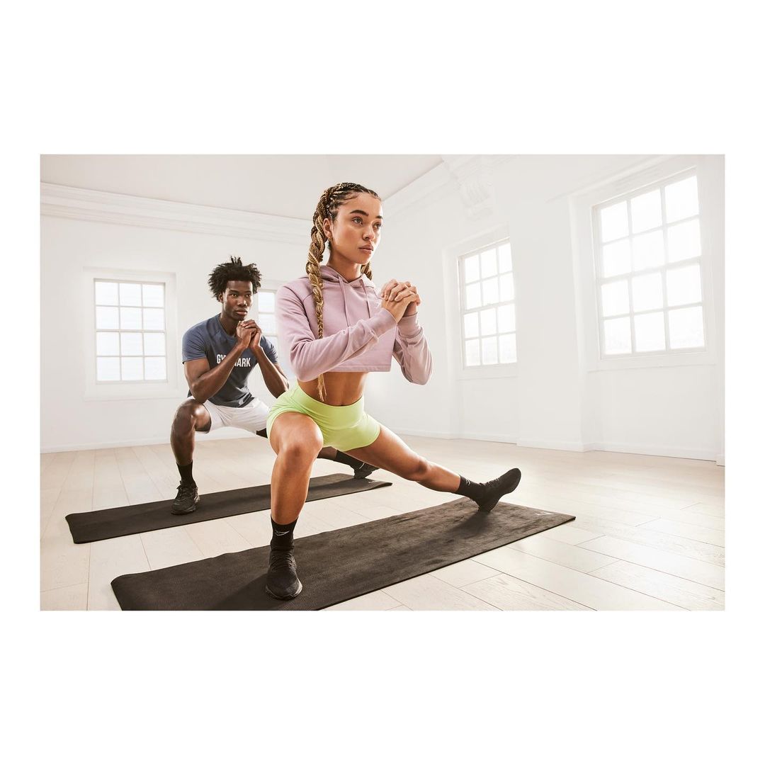 Athletic man and woman in activewear perform side lunges on black exercise mats in a bright, white-walled fitness studio.