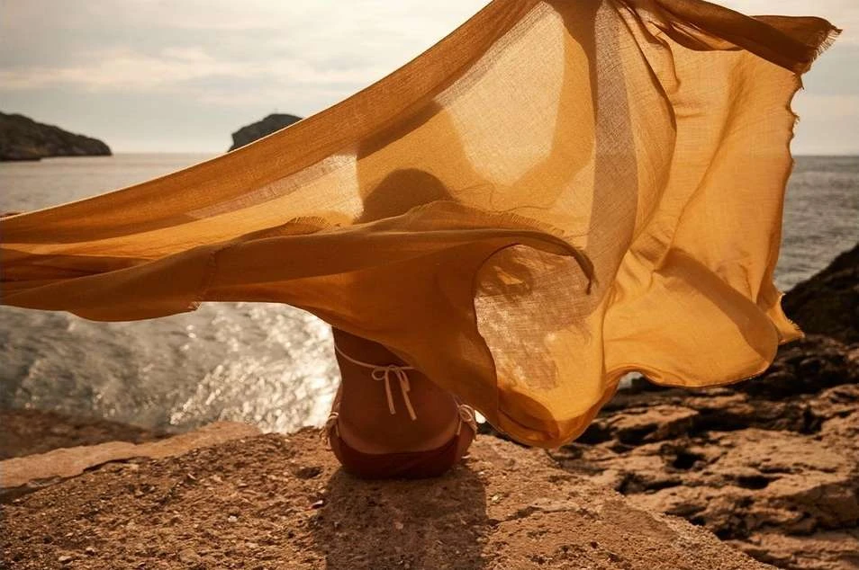 Woman in swimwear sitting on rocky shore near sea, holding up flowing golden fabric in wind, with island in background.