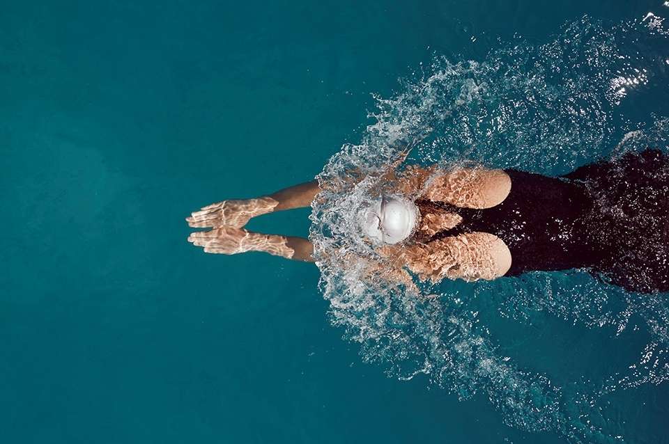 Overhead shot of a swimmer with a white cap in a turquoise pool, creating splashes during the butterfly stroke, captured in an eye-level angle.